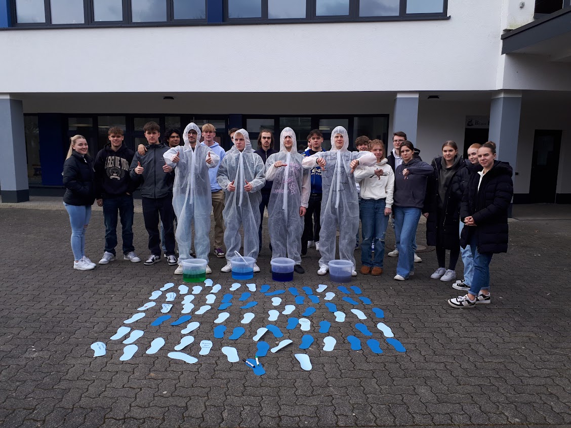 Schüler:innen positionieren sich vor vier Eimern, die mit verschiedenfarbigem Wasser gefüllt sind, sowie vor zahlreichen unterschiedlich farbigen Fußabdrücken.